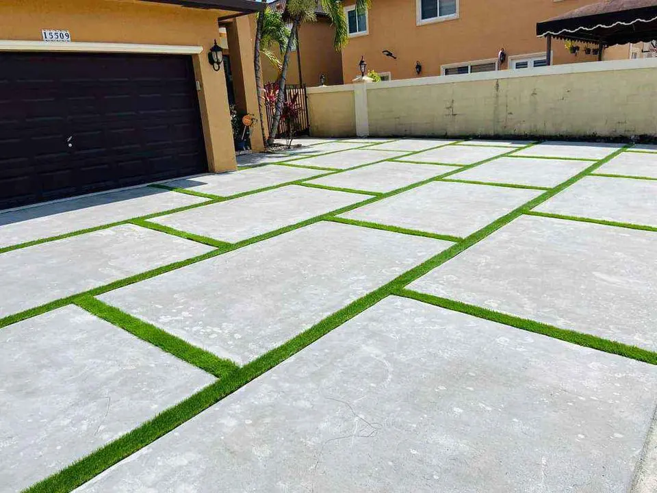 A driveway with large square concrete slabs separated by strips of lush ABQ Turf, leading to a dark garage door. Beige house walls with windows are visible in the background.