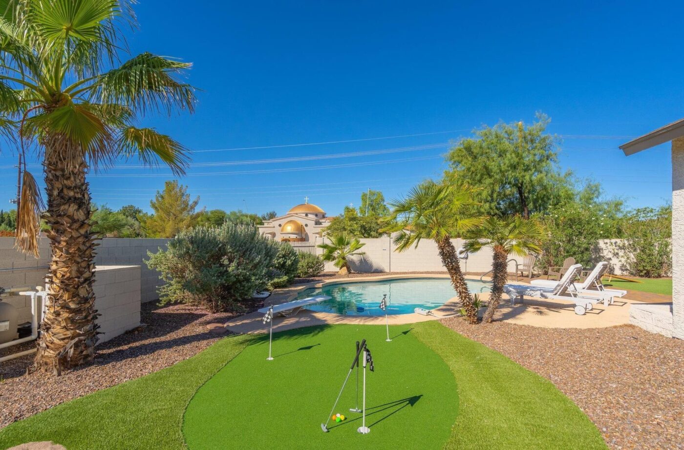 Nestled in a Santa Fe, NM backyard, a small putting green with artificial grass is bordered by palm trees. A round swimming pool is encircled by lounge chairs under the clear blue sky, with a stucco wall and building in the distance. This serene oasis offers ideal low-maintenance solutions.
