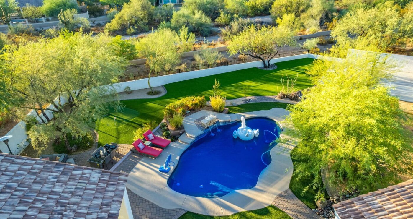 A backyard featuring ABQ Turf's lush synthetic turf, a circular stone fire pit at its heart, surrounded by mature trees and plants. A stone border lines the immaculate grass, all under a clear blue sky.