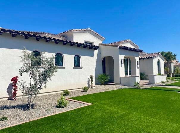 A modern white stucco house with a terracotta tiled roof. The front features arched windows and a small porch. The landscape, enhanced by pet turf for low-maintenance solutions, includes a green lawn, gravel, and small plants under a clear blue sky.