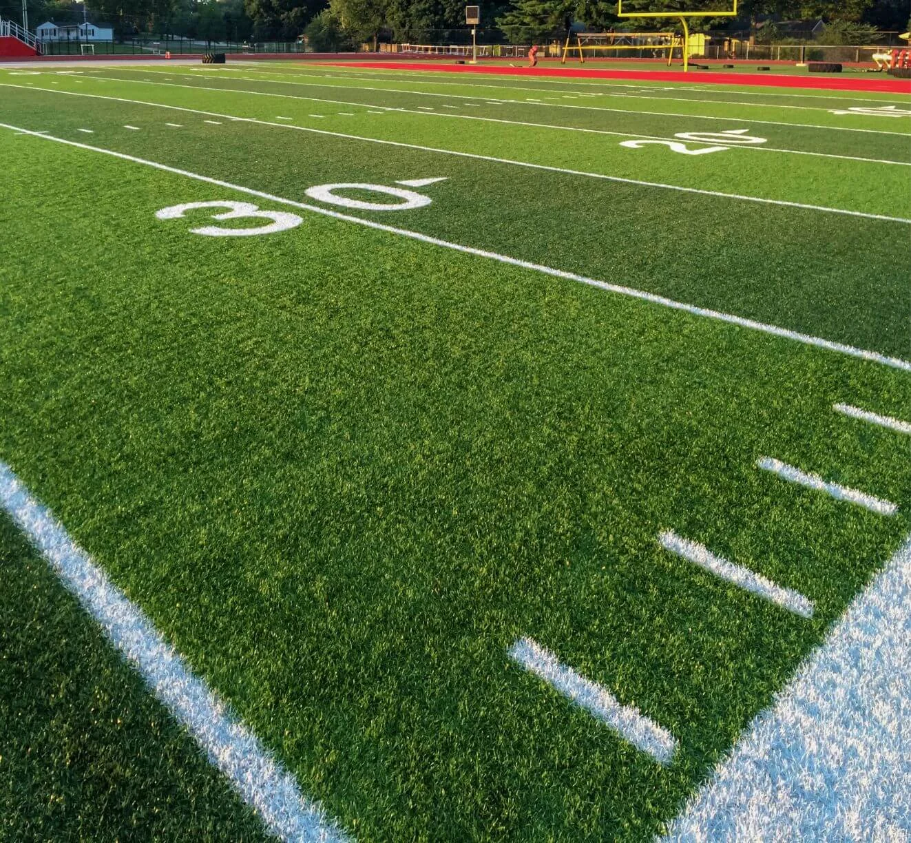 View of an American football field with vibrant green turf installed by expert artificial grass installers, showing the 30-yard line marked in white. In the background, there's a yellow goalpost, a running track, and trees lining the stadium. The sunlit scene captures a perfect day for play.