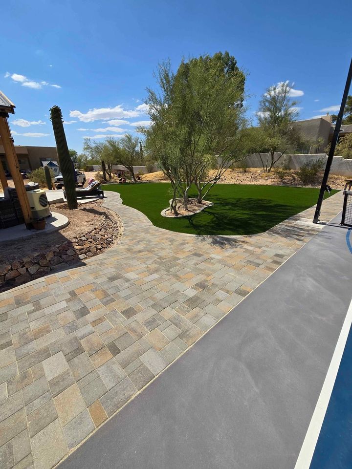 A backyard with a stone pathway leads to a grassy area adorned with desert plants and trees, while a basketball court adds an active element. In the foreground, brick patios hint at expert craftsmanship akin to Santa Fe masonry, against a backdrop of clear blue sky and desert expanse.
