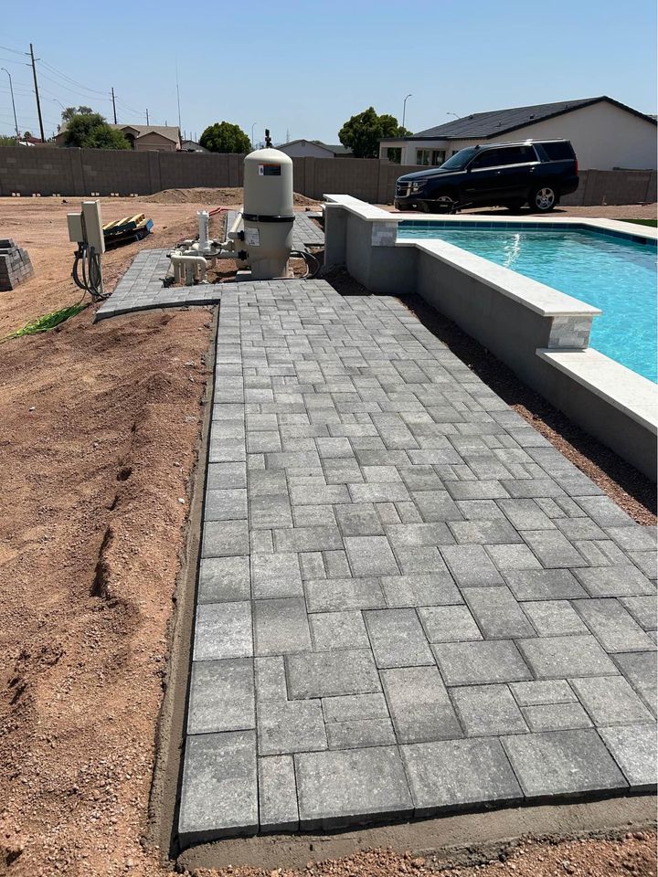 A newly constructed pool area boasts elegant gray stone tiles installed by expert paver installers. A black car rests beside it, amid unfinished landscaping and dirt piles. Nearby, a filter system hums quietly. Under the clear blue sky, this scene promises future transformation into a Santa Fe masonry masterpiece.
