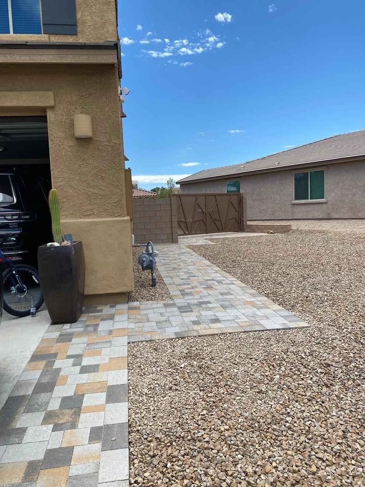 A house exterior displays a paved walkway flanked by a small cactus in a planter, leading up to the driveway. The yard, with its gravel cover, embraces a Santa Fe masonry vibe under the clear, blue sky.