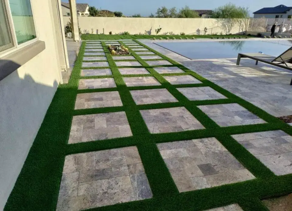 A modern outdoor patio with a checkered design, featuring large square stone tiles and pet-friendly turf accents. The area leads to a swimming pool with lounge chairs, surrounded by a garden with small plants and trees in the background.